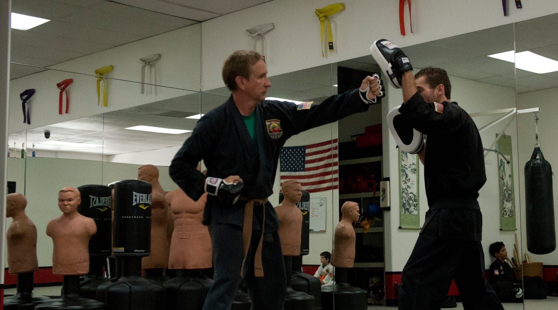 An instructor holding a focus pad for an adult student practicing strikes at USSD Santa Barbara, with the belt rank display on the wall behind them.