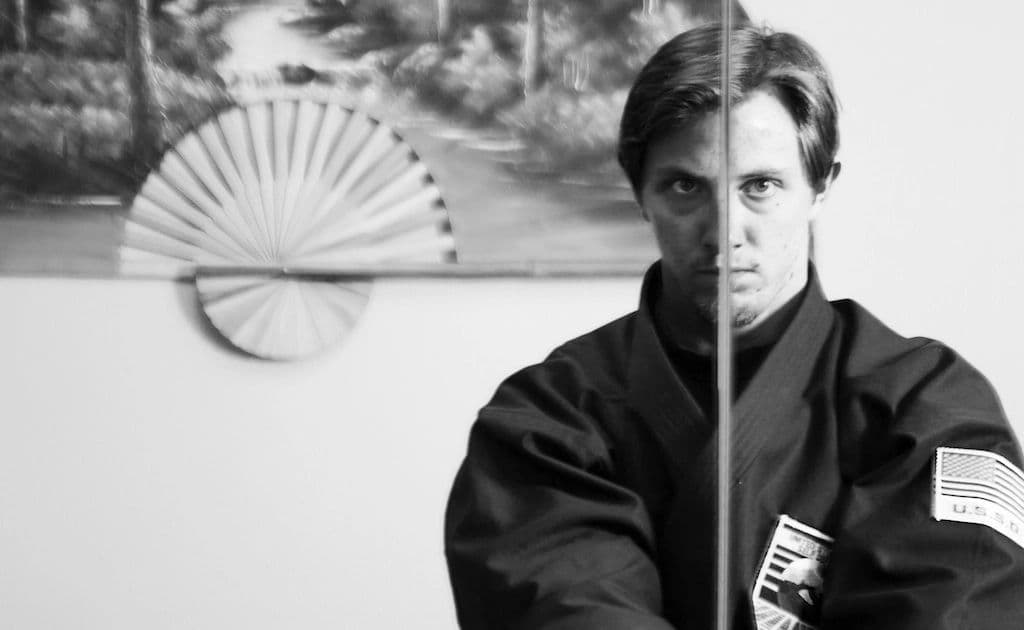 Man in martial arts uniform holding a sword, split face reflection.