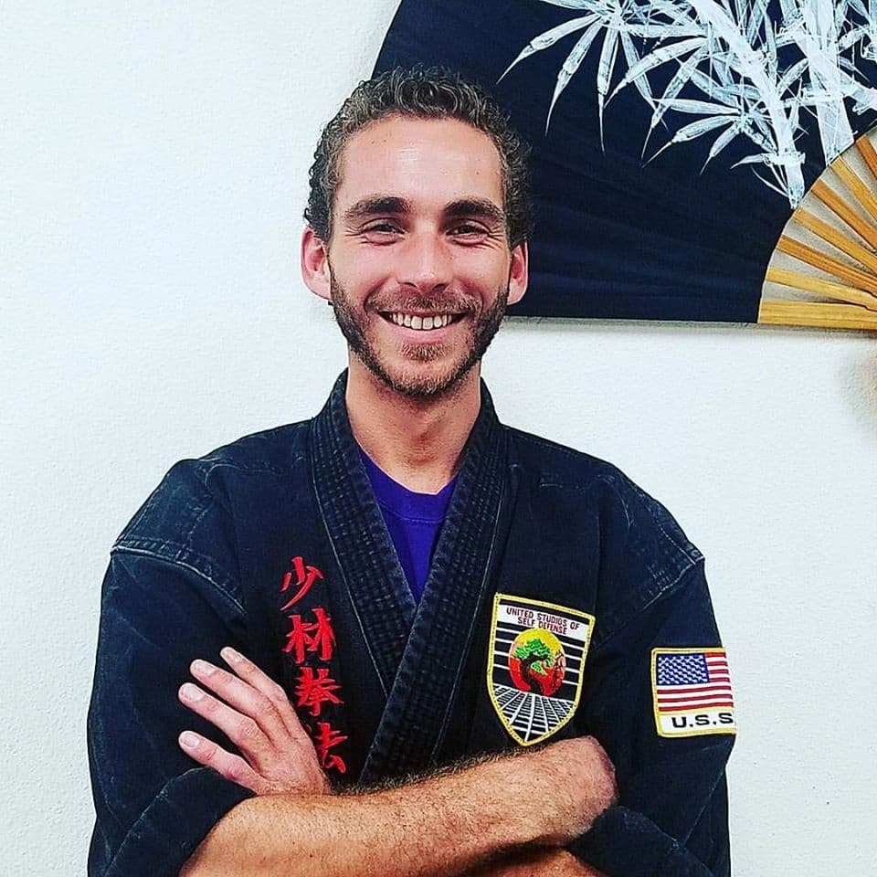 Smiling martial artist in a black gi with patches and folded arms.